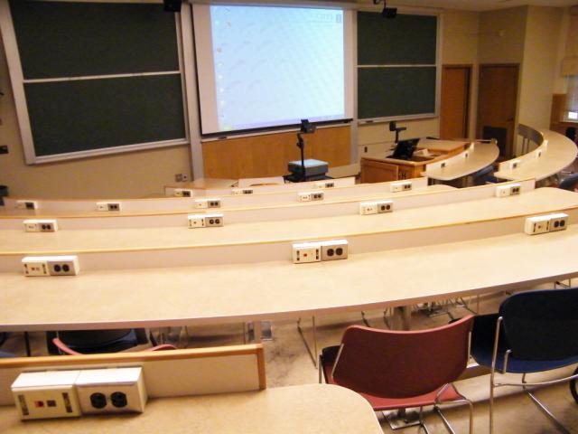 A view of the classroom with tiered sloped seating, chalkboard, and instructor table in front.
