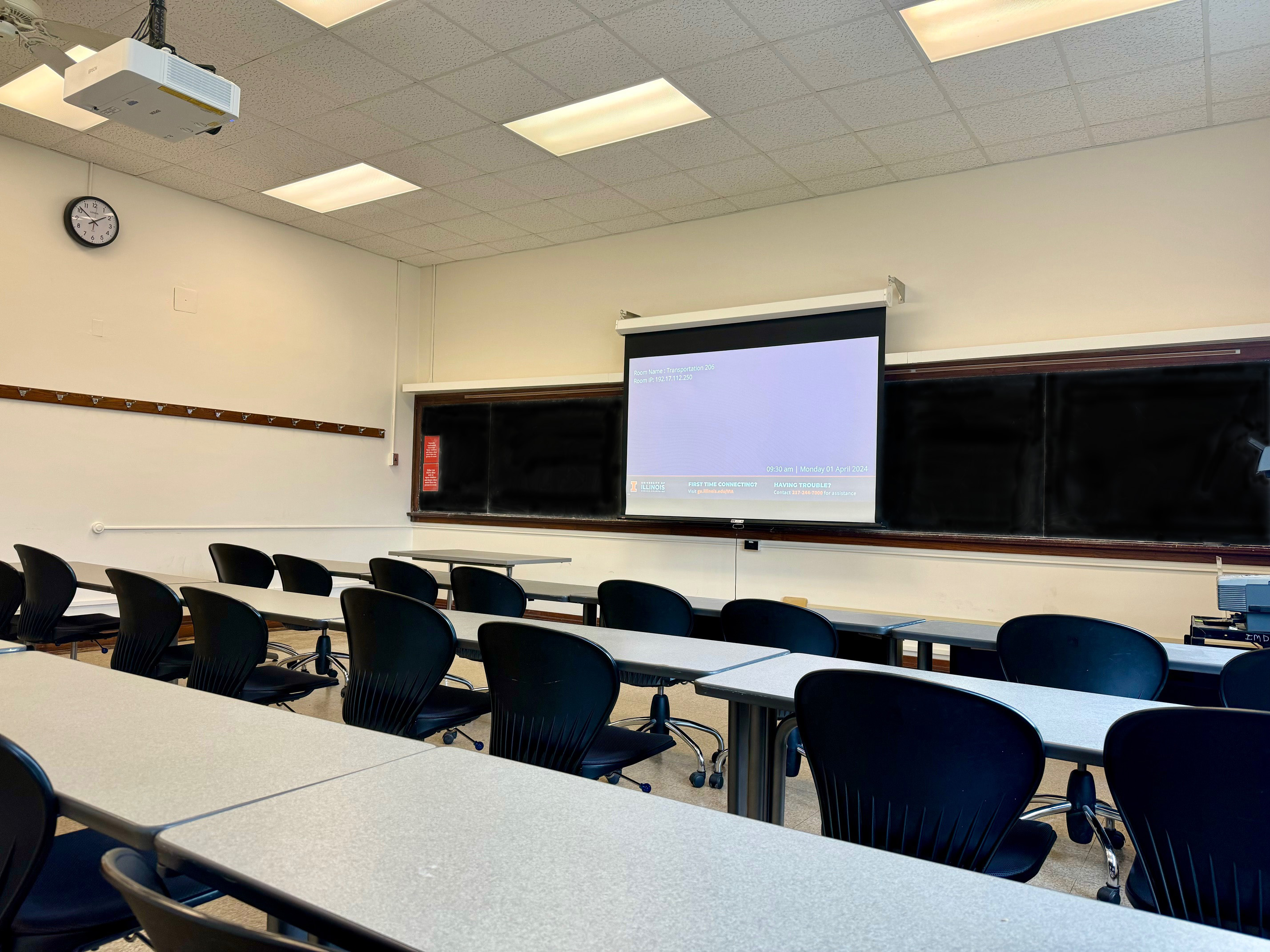 A view of the classroom with student desks, a front lecture table, and chalkboard.