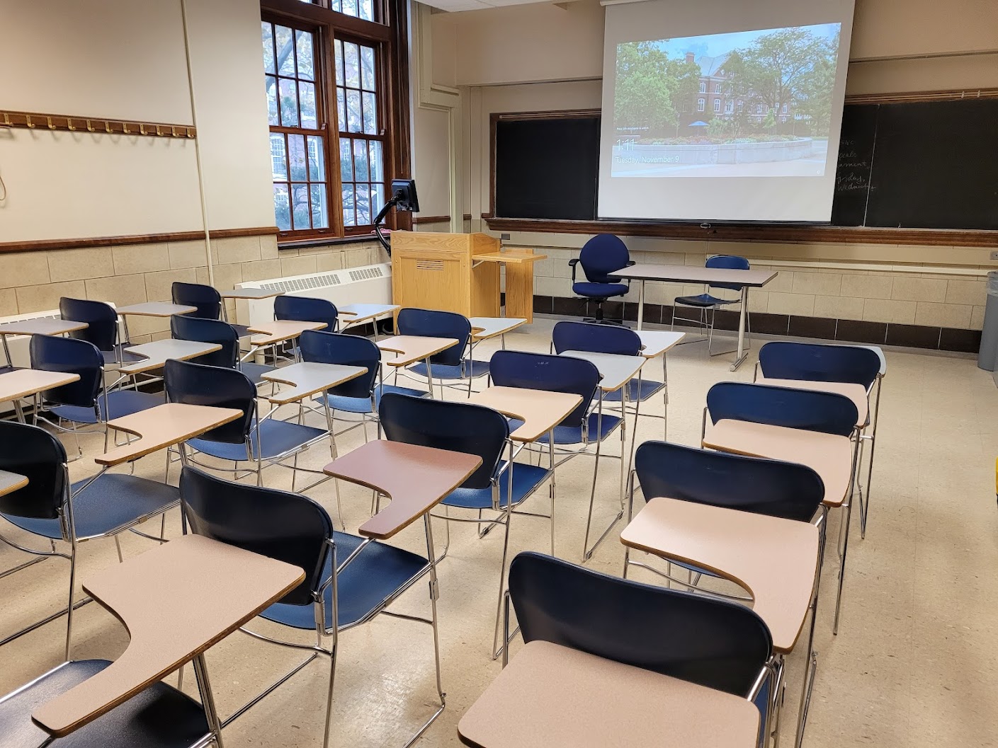 A view of the classroom with movable tablet arm chairs, chalkboard, and instructor table in front.