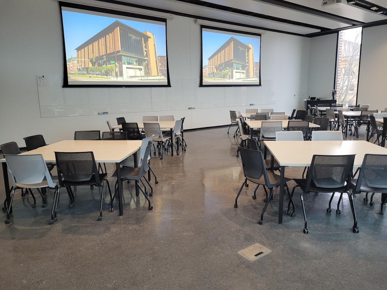 This is a view of the room with student tables and chairs, projection screens, a front lecture table, glass boards, and windows.