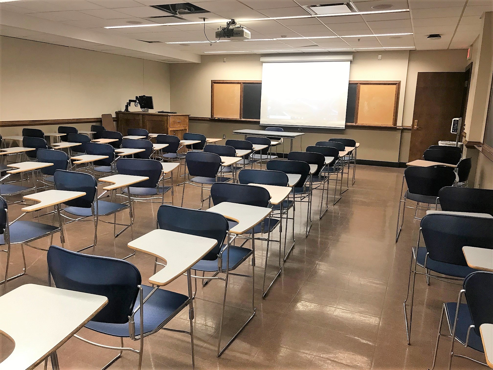 A view of the classroom with movable tableted arm chairs, chalkboard, and instructor table in front. 