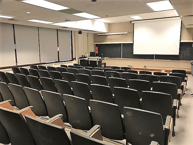 A view of the classroom with theater auditorium seating, chalkboard, projection screen, instructor lectern and table in front.