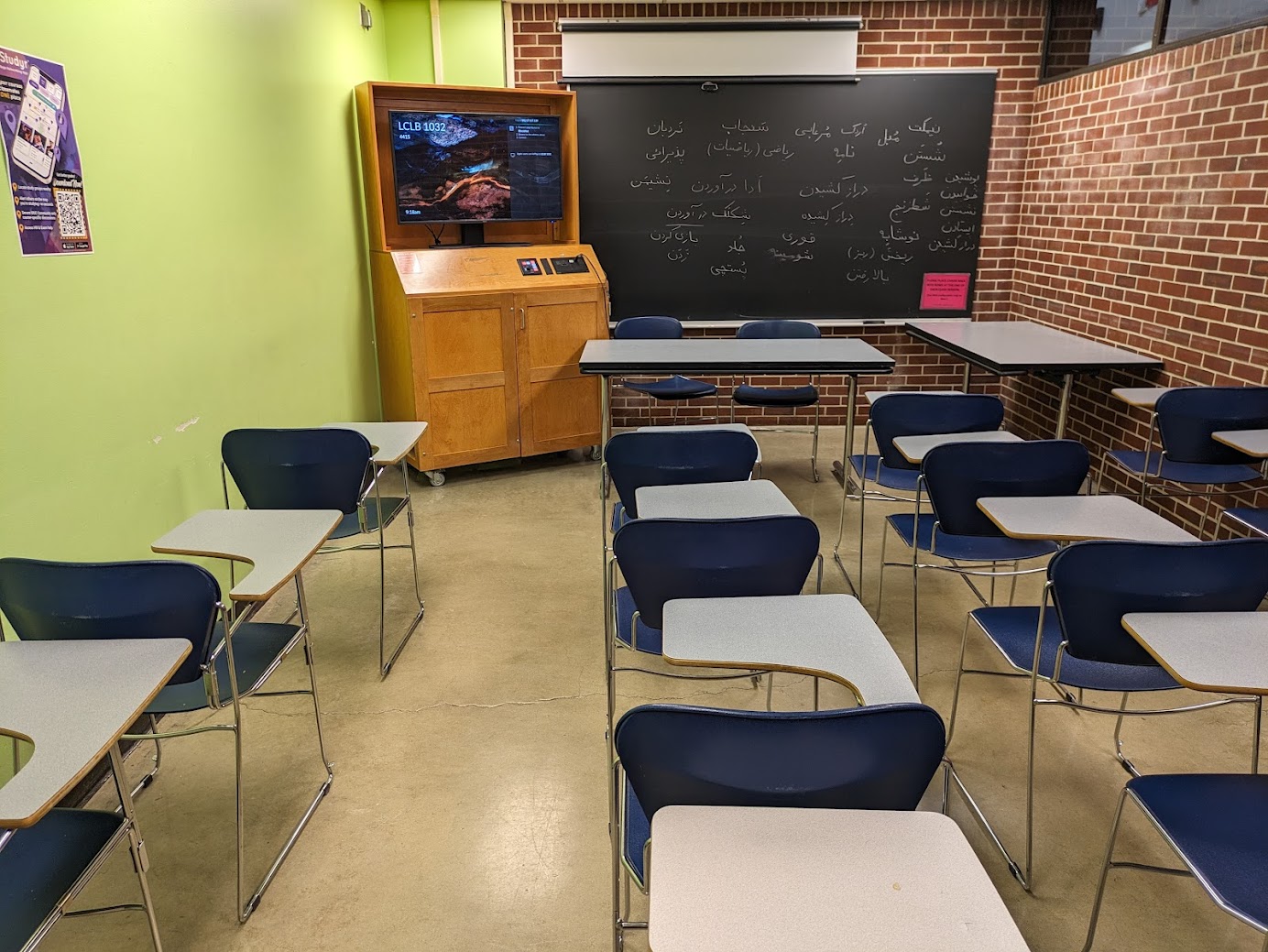 A view of the classroom with movable tableted arm chairs, chalk board, LCD monitor in cabinet, and instructor table in front.