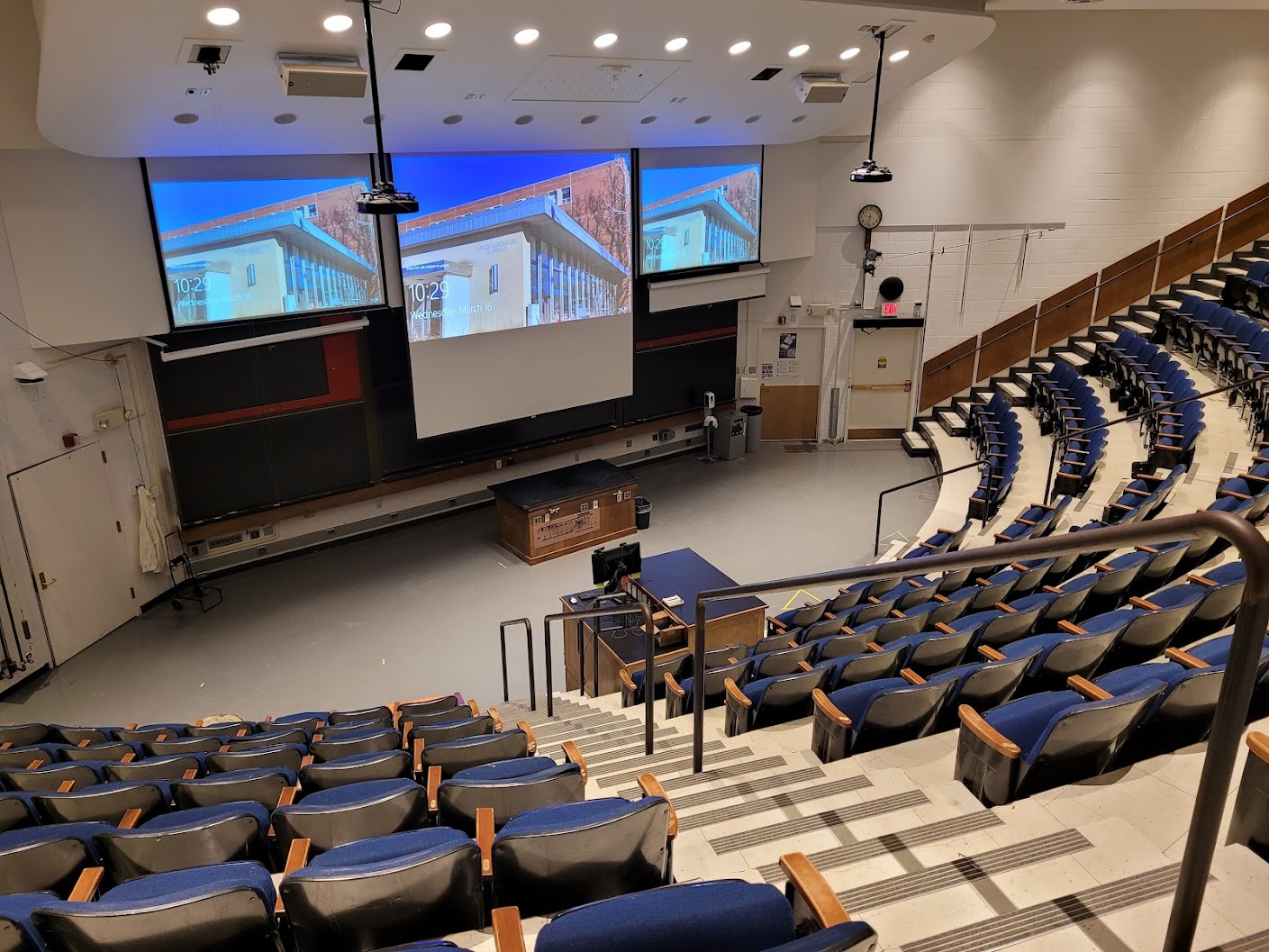 A view of the classroom with theater auditorium seating, chalk boards, projection screens, and instructor cabinet in front.