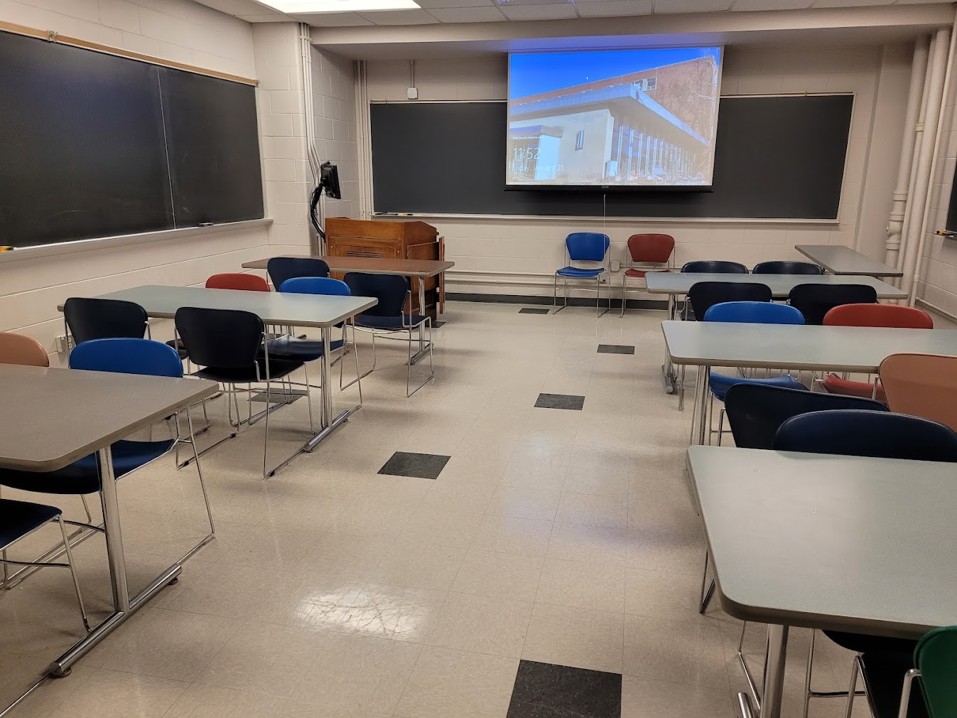 A view of the classroom with movable tables and chairs, chalkboard, and instructor table in front.