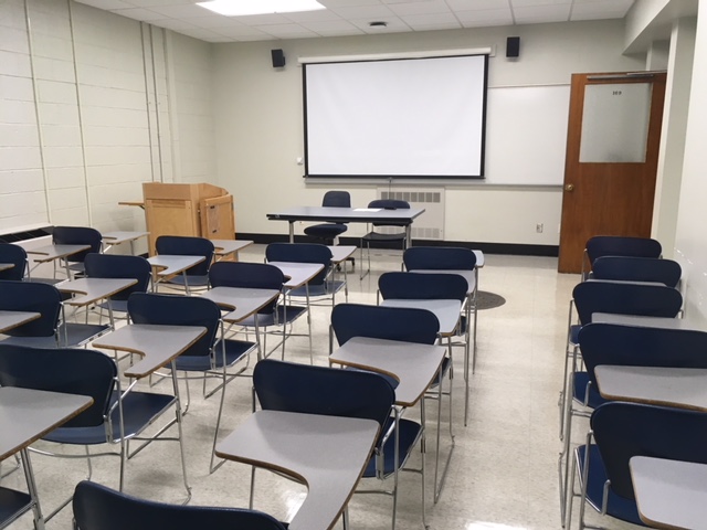 A view of the classroom with movable tableted arm chairs, chalkboard, and instructor table in front.