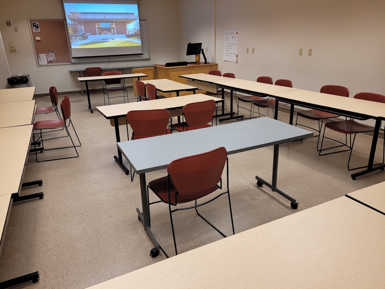 A view of the classroom with moveable tables and chairs, whiteboard, projection screen, and instructor table in front.
