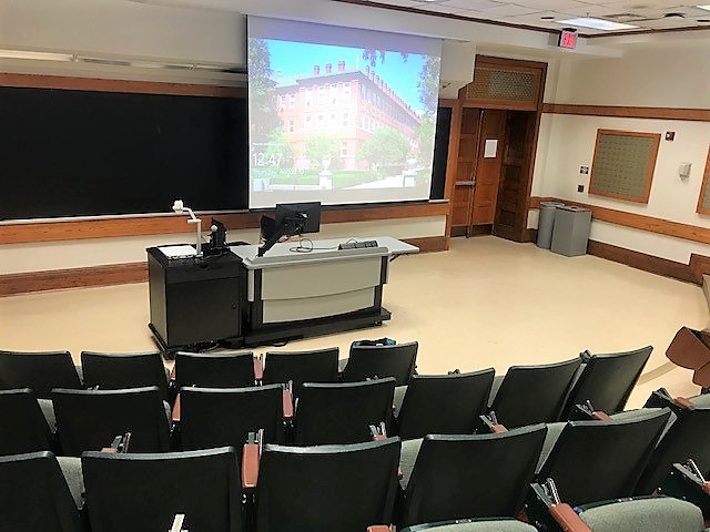 A view of the classroom with theater auditorium seating, projector and screen, chalkboard, instructor lectern, and instructor table in front.