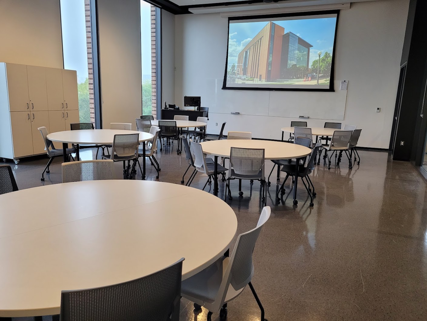 This is a view of the room with student desks, a front lecture table, and a glass board.