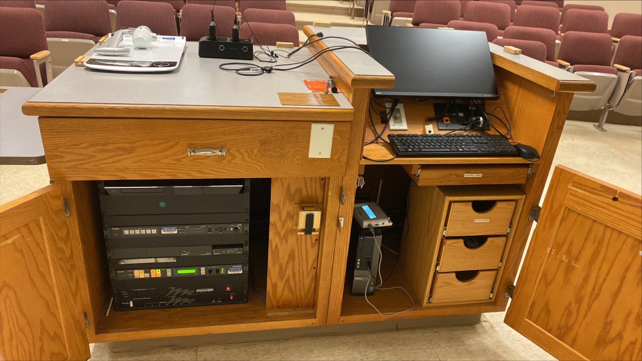 A view of the open lectern with computer monitor, PC, Document Camera, and Blu-ray, a push button controller, and audio-visual rack.