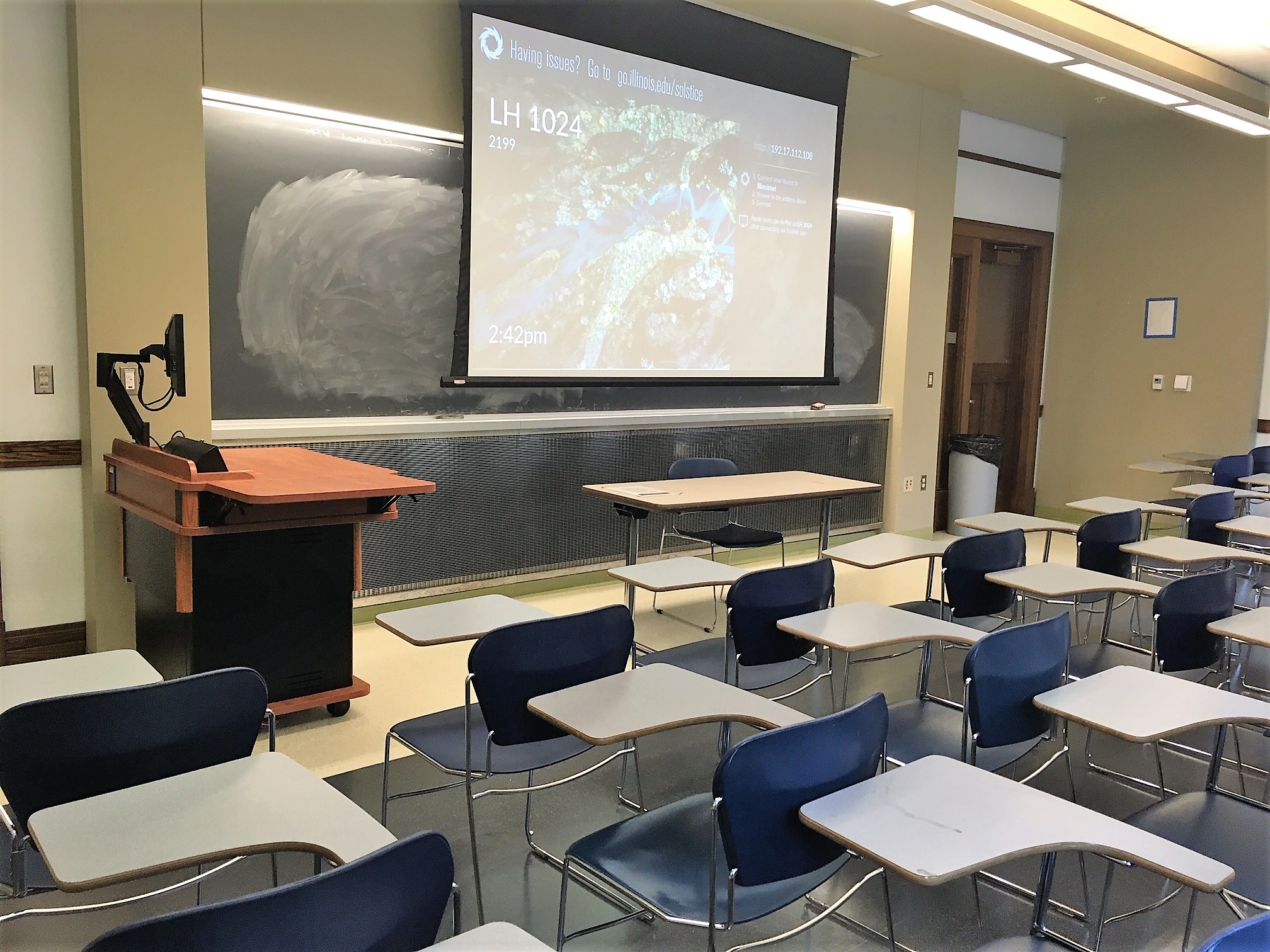 A view of the classroom with moveable tablet arm chairs, chalkboard, and instructor table in front.