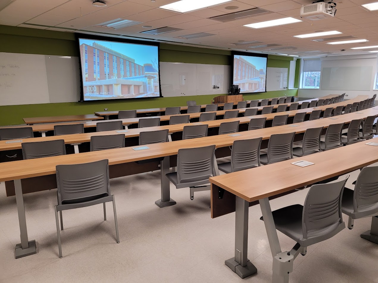 A view of the classroom with fixed tables and chairs, white boards, and instructor table in front