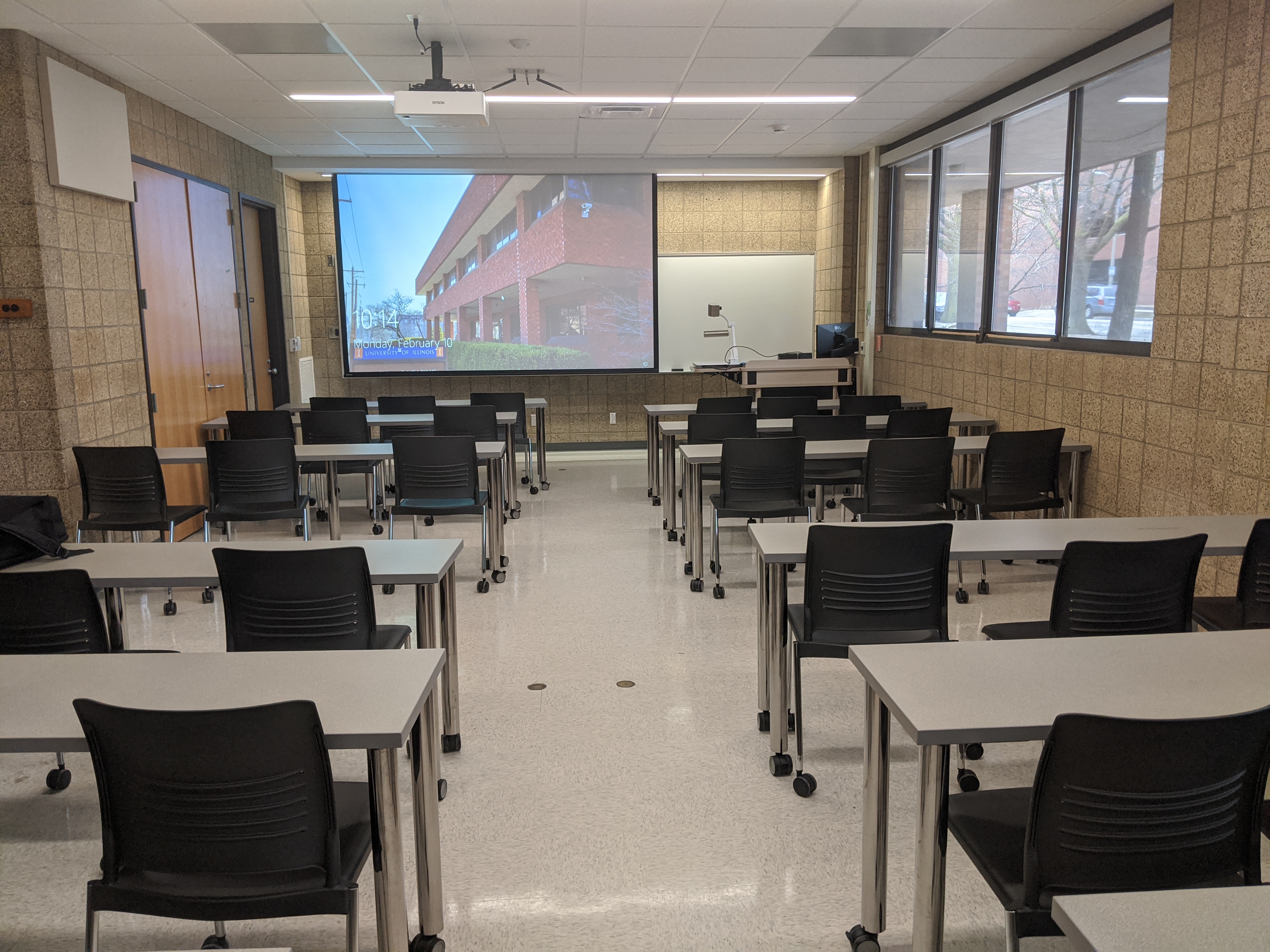 A view of the classroom with movable tables and chairs, a front instructor lectern, projection screen, and white board.