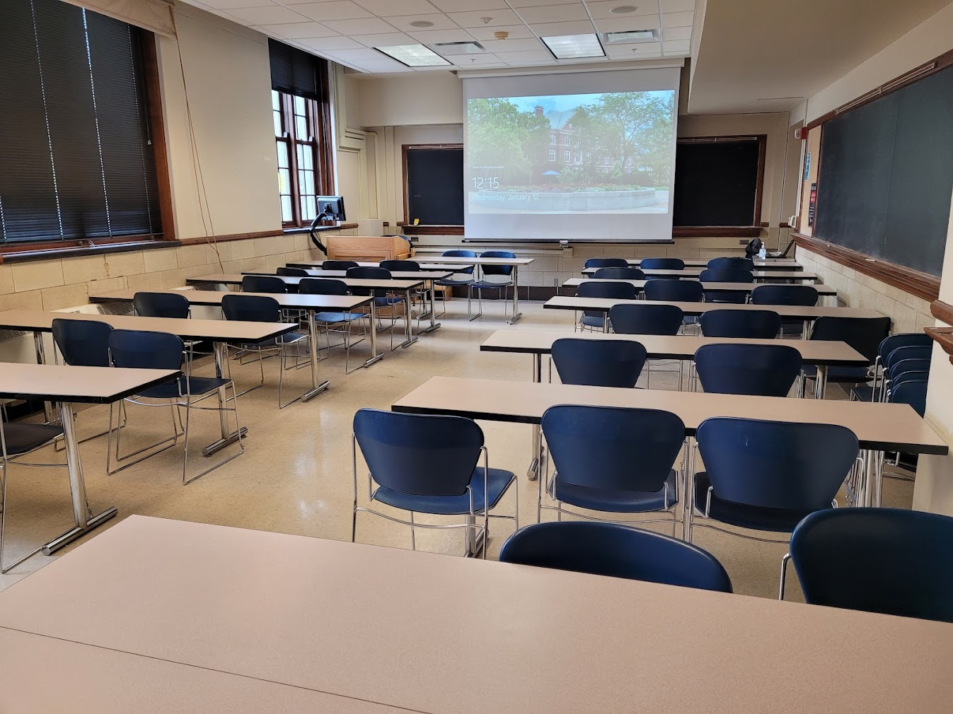 A view of the classroom with moveable tables and chairs, chalkboard, and instructor table in front.