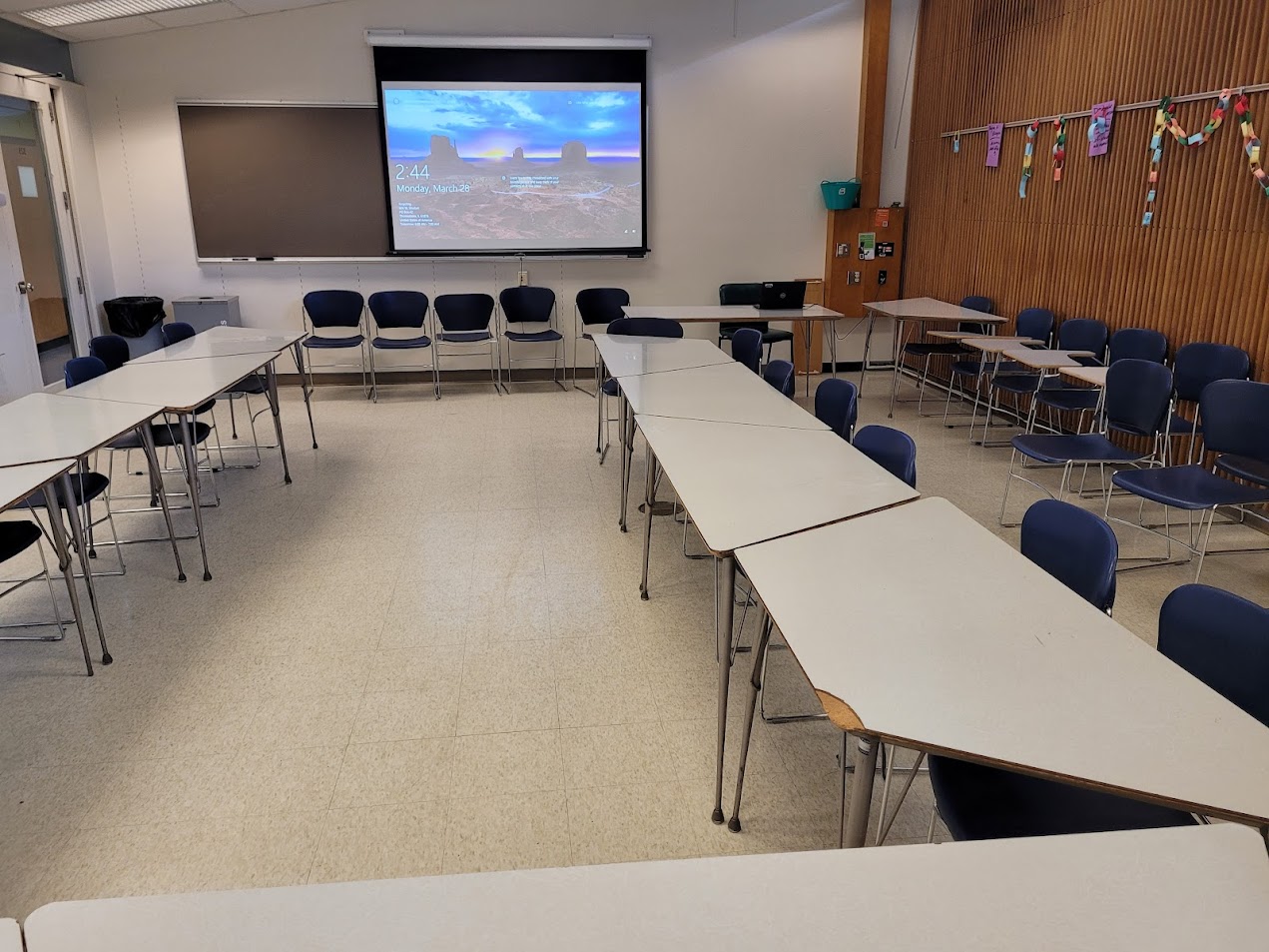 A view of the classroom with moveable tables and chairs, chalkboard, and instructor table in front