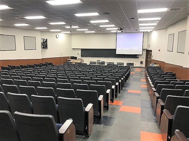 A view of the classroom with theater auditorium seating, chalkboard, and instructor table in front.