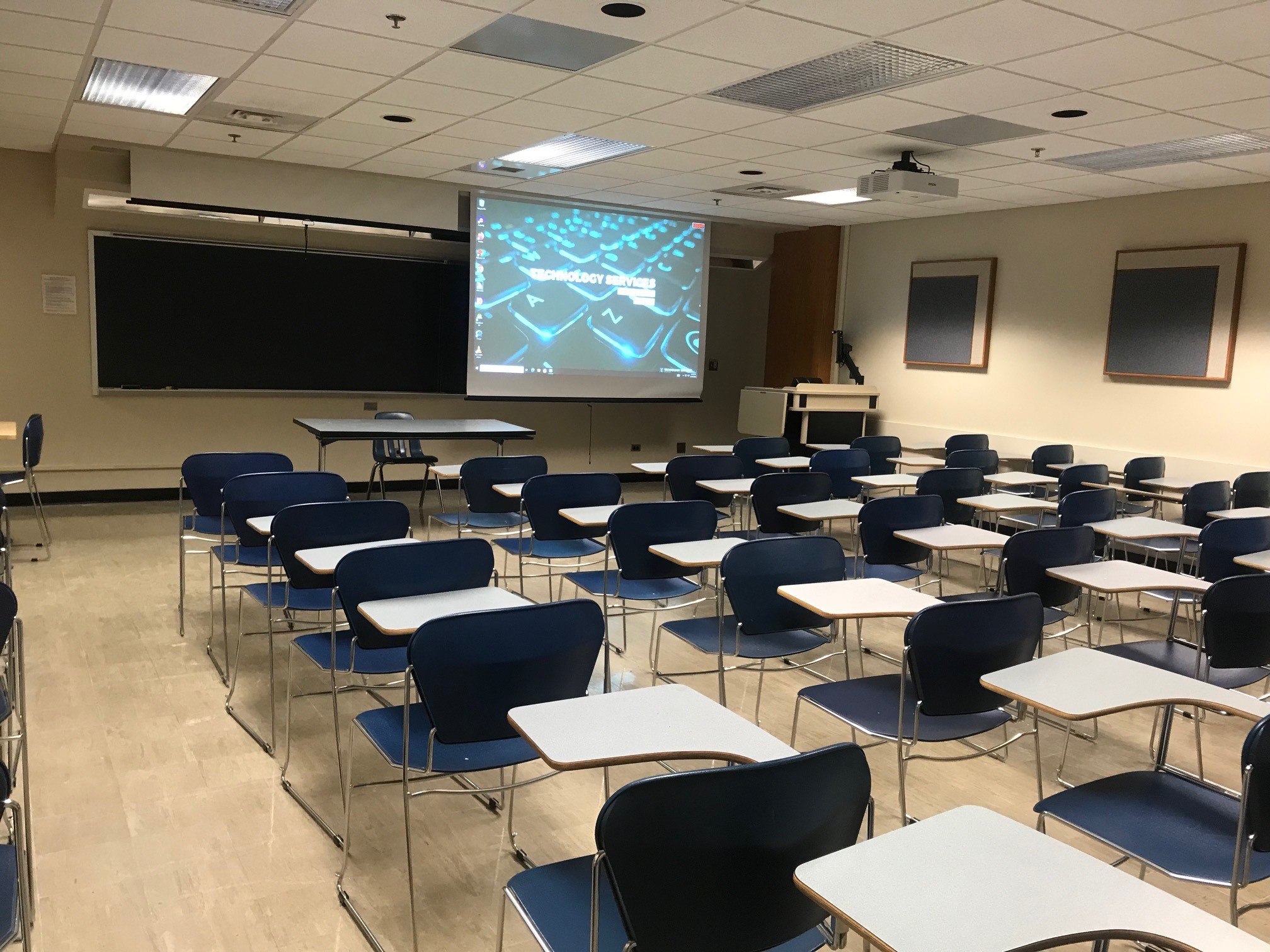 A view of the classroom with movable tableted arm chairs, chalkboard, instructor lectern, and instructor table in front.