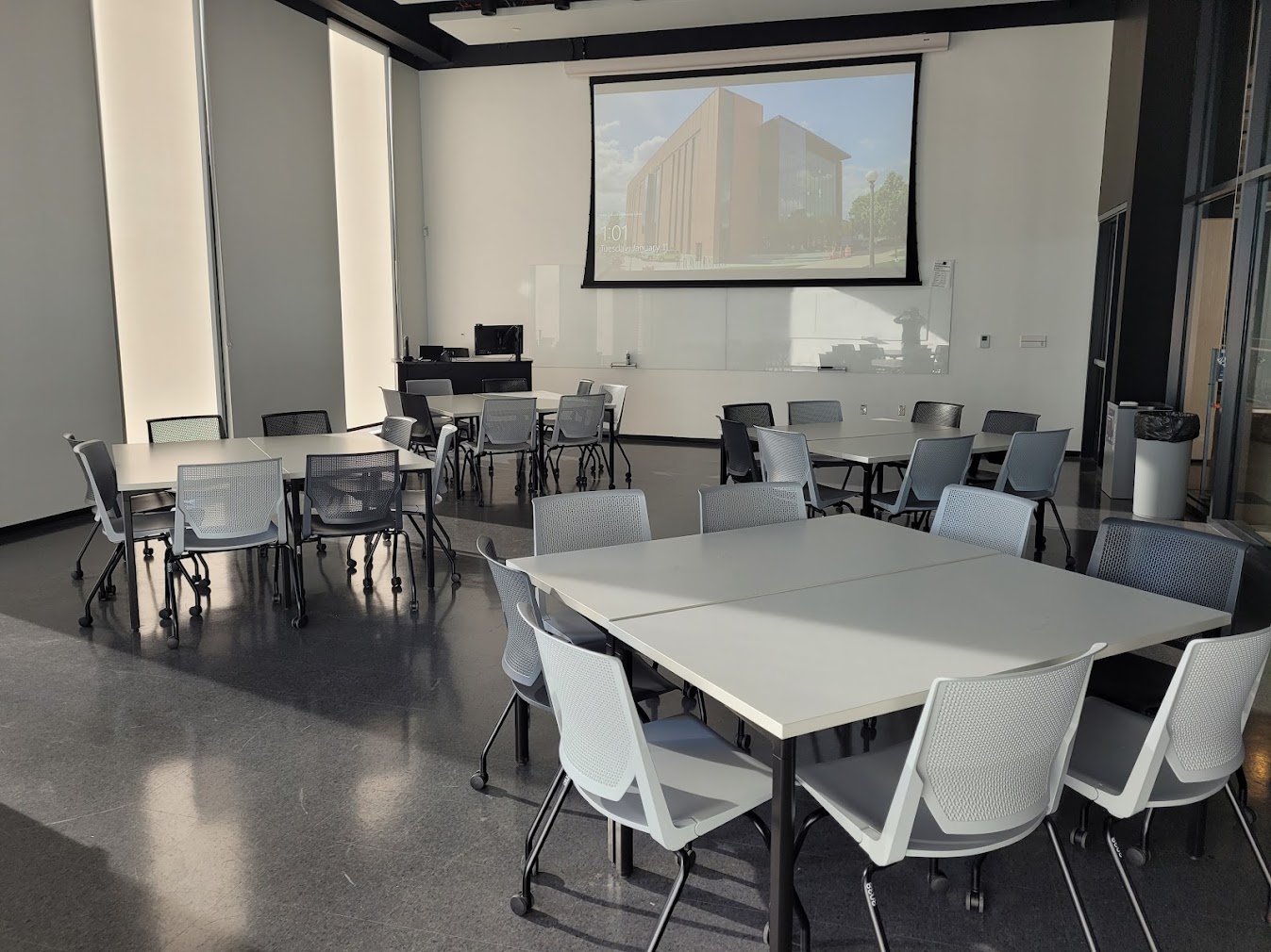 This is a view of the room with student desks, projection screen, a front lecture table, windows, and a glass board.