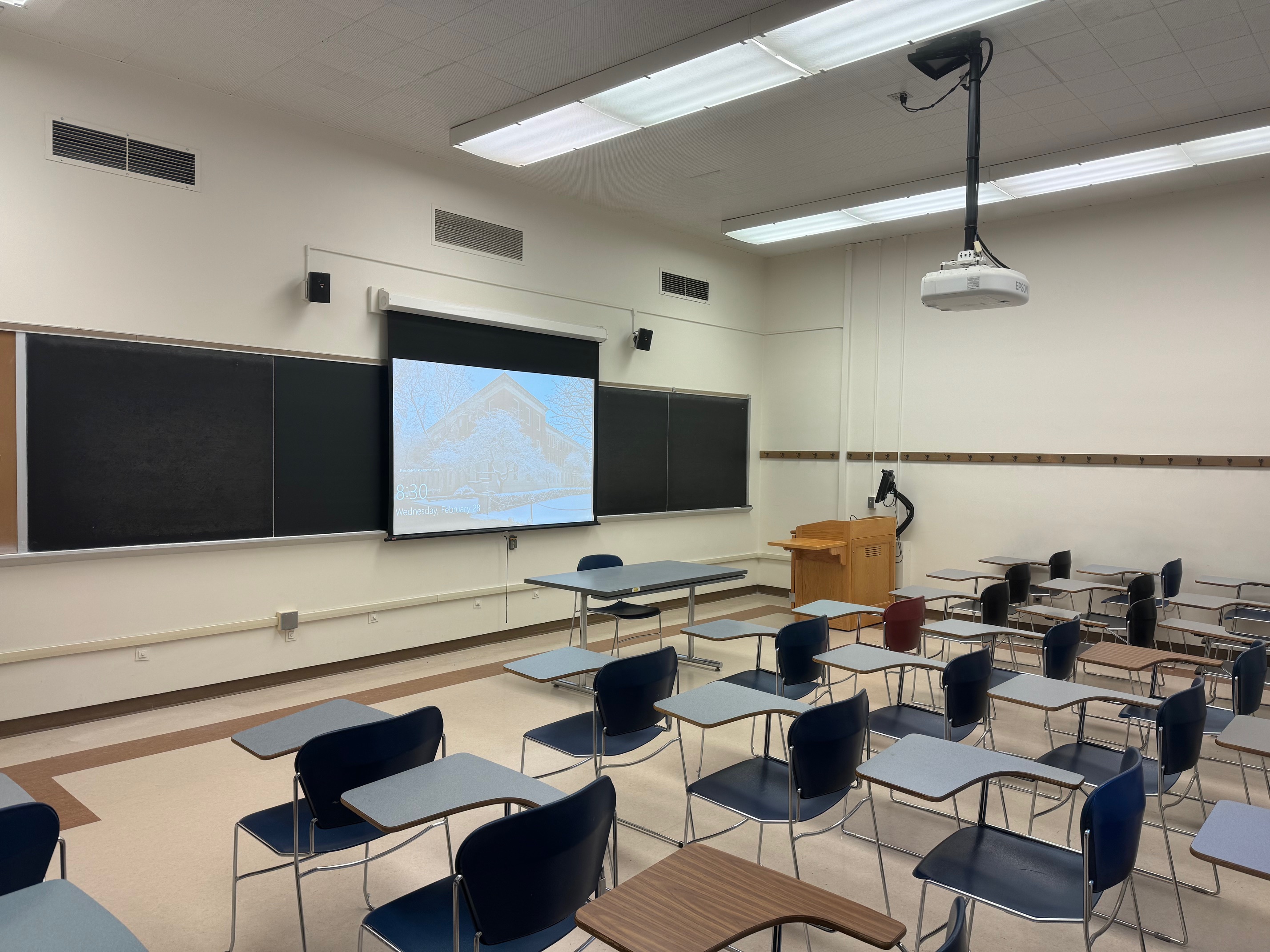 A view of the classroom with Movable Tableted Arm Chairs, projector and screen, chalkboard, instructor lectern, and instructor table in front.