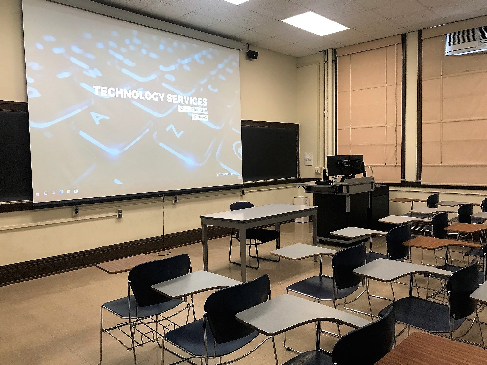 This is a view of the room, with student desks, a front lecture table, projection screen, instructor table, and a chalkboard.