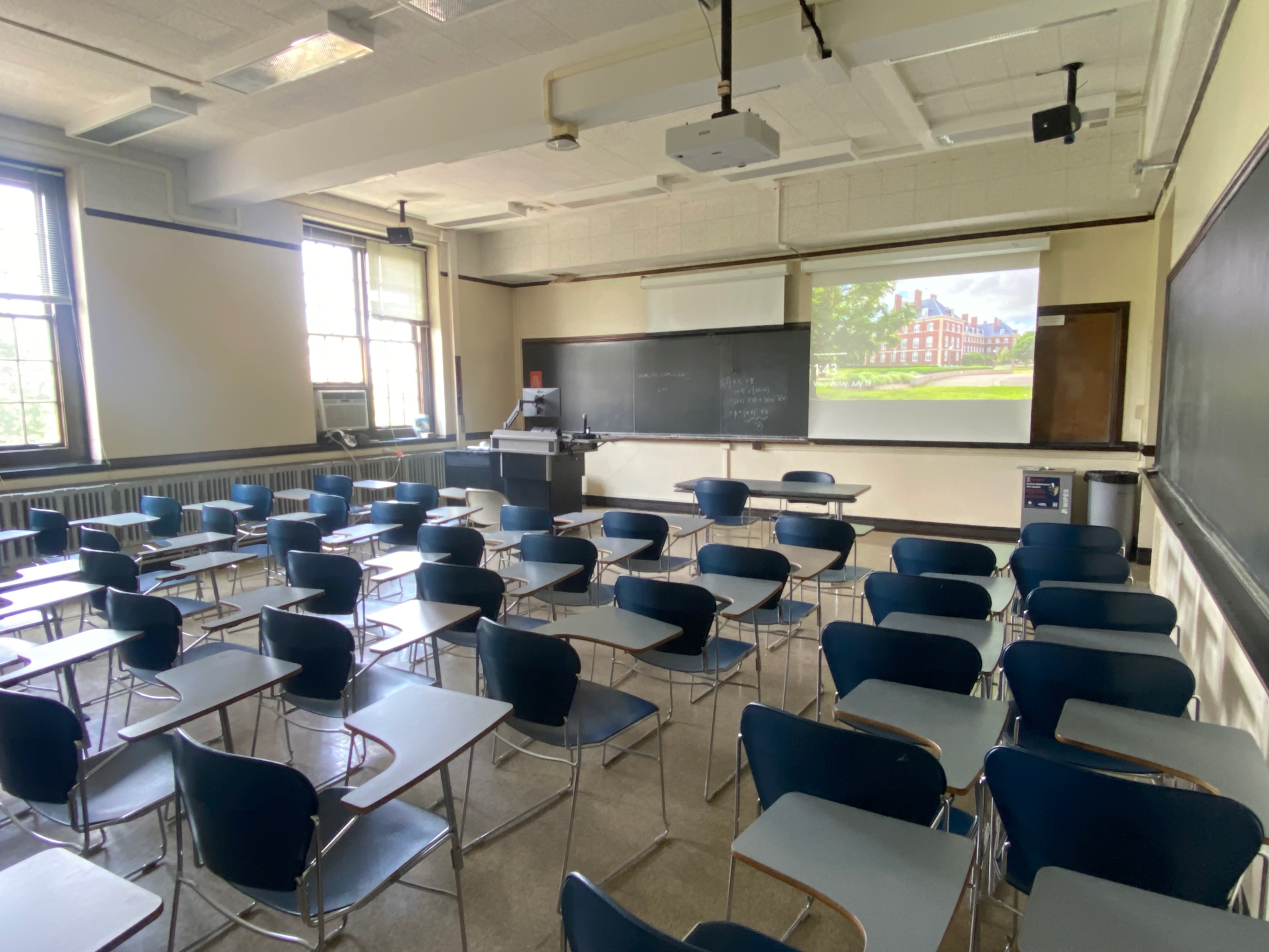 A view of the classroom with Moveable Tableted Arm Chairs, projector and screen, chalkboard, instructor lectern, and instructor table in front.