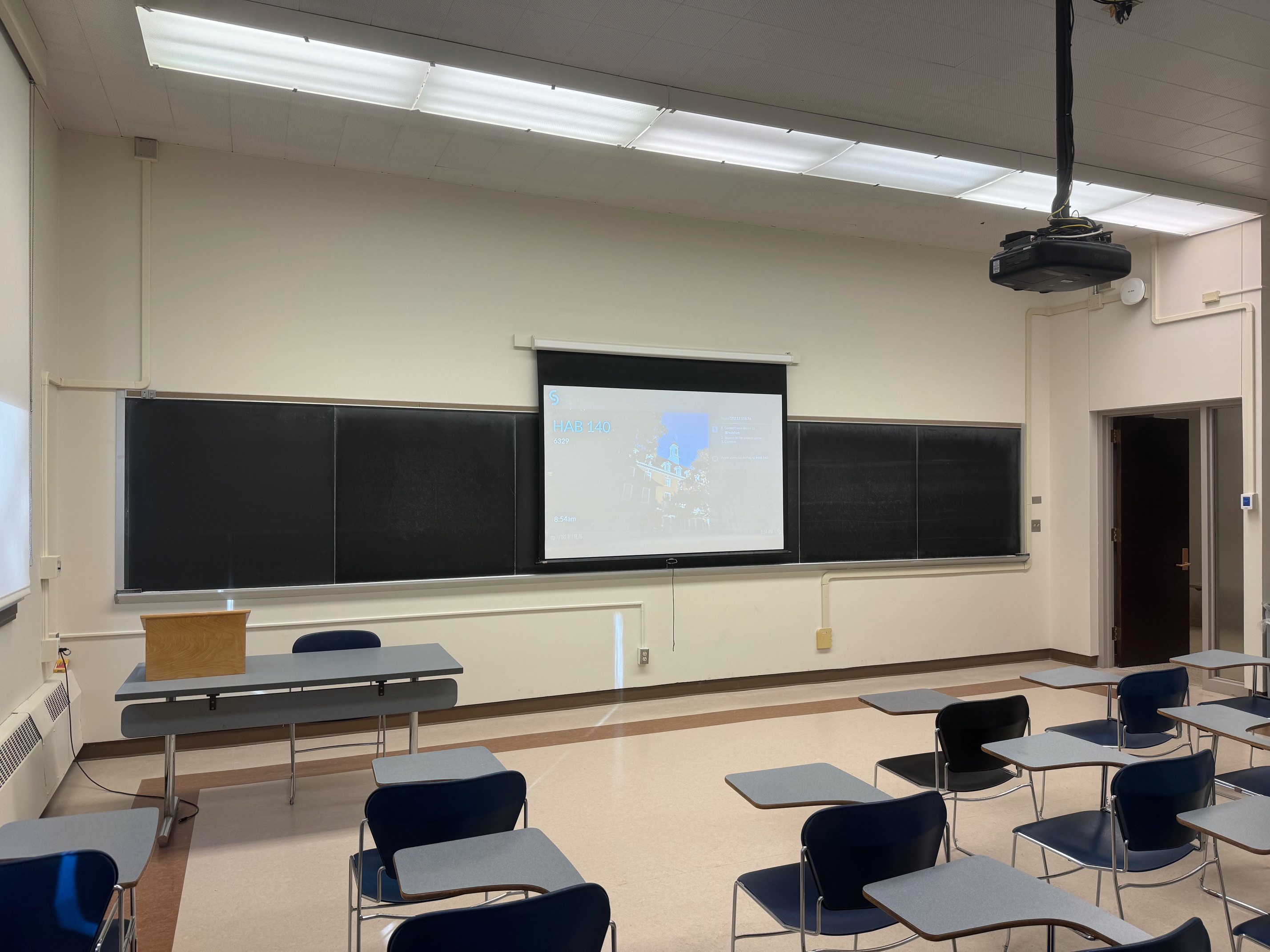 A view of the classroom with movable tableted arm chairs, instructor table, projection screen and chalkboard.