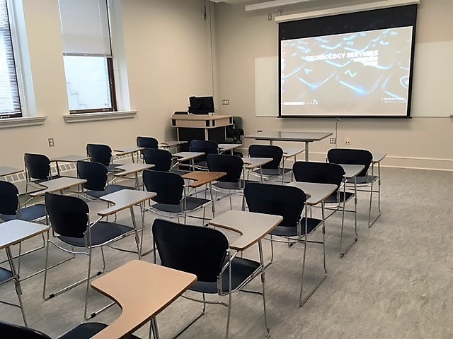 A view of the classroom with movable tableted arm chairs, white board, and instructor table in front.