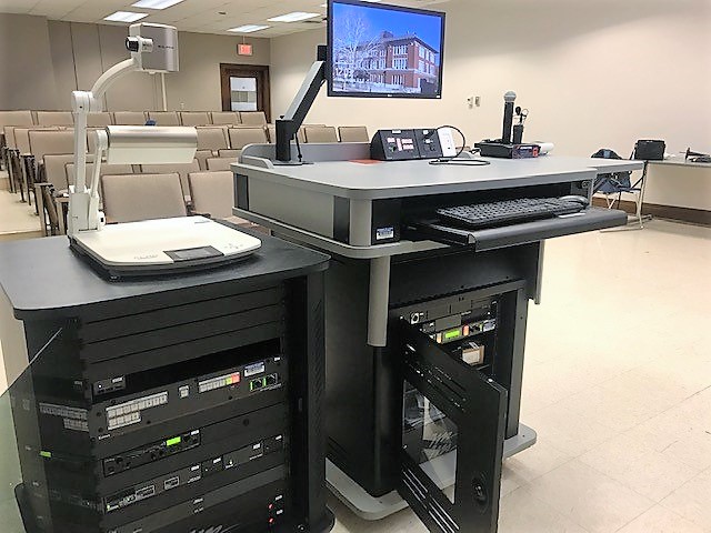 A view of the open cabinet with a computer monitor, HDMI inputs, a push button controller, document camera, microphones, audio visual rack, and electric screen controls.