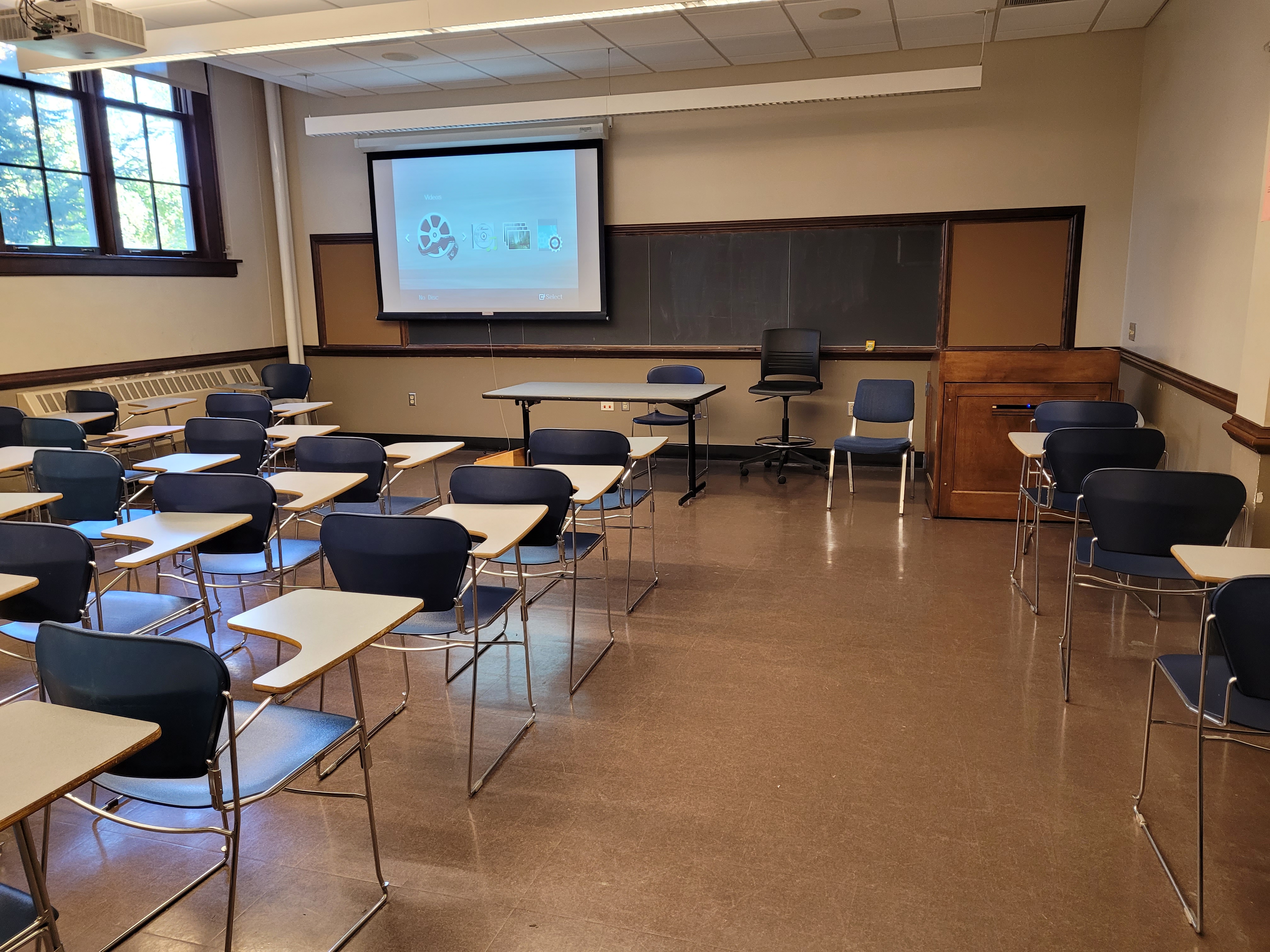 A view of the classroom with movable tableted arm chairs, chalk board, and instructor table in front.