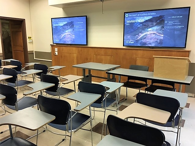 A view of the classroom with movable tablet arm chairs, LCD wall monitors, and instructor table in front
