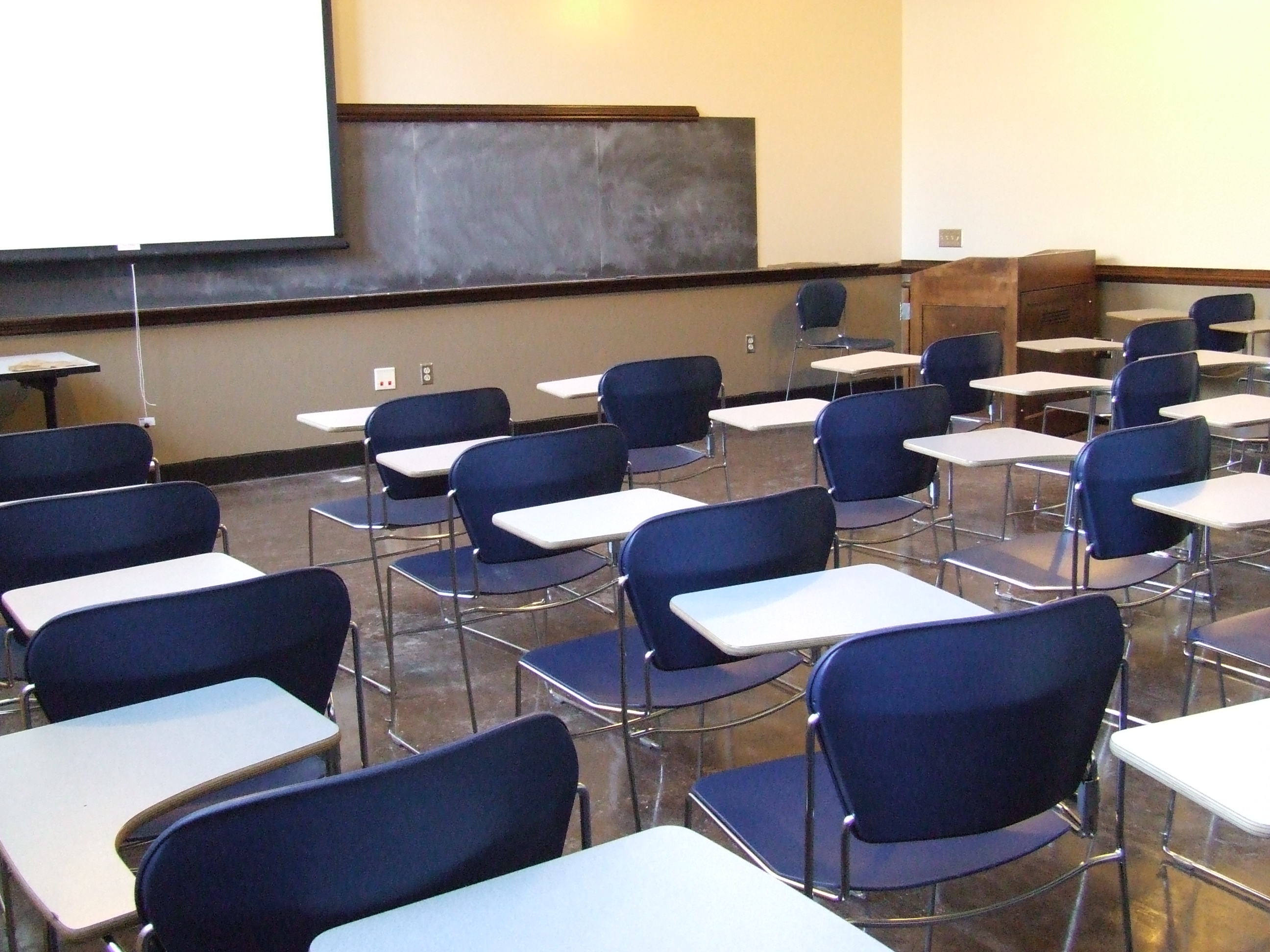A view of the classroom with movable tableted arm chairs, chalkboard, and instructor table in front.