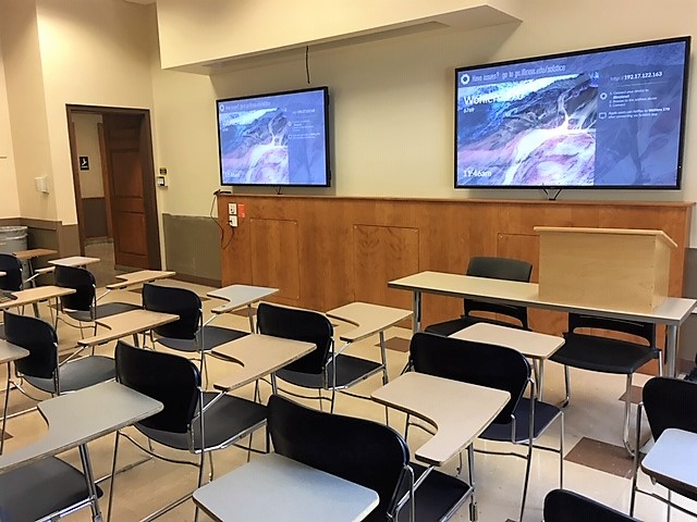 A view of the classroom with movable tablet arm chairs, wall monitors, and instructor table in front.