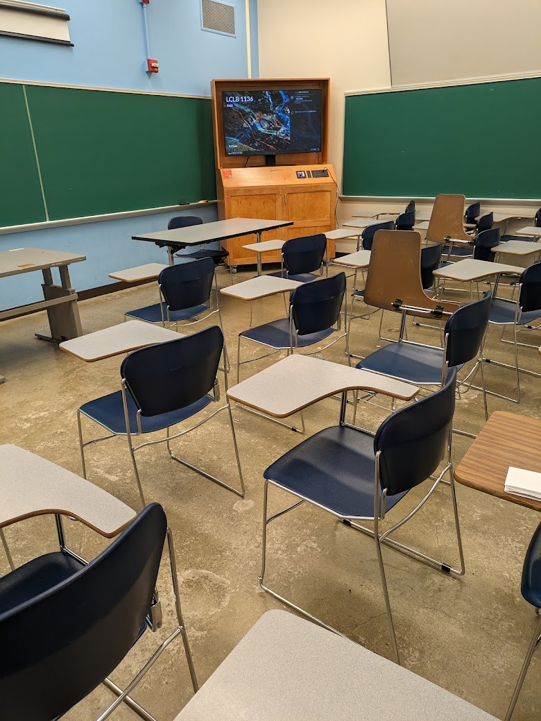 A view of the classroom with movable tableted arm chairs, chalkboard, and an instructor table in front.