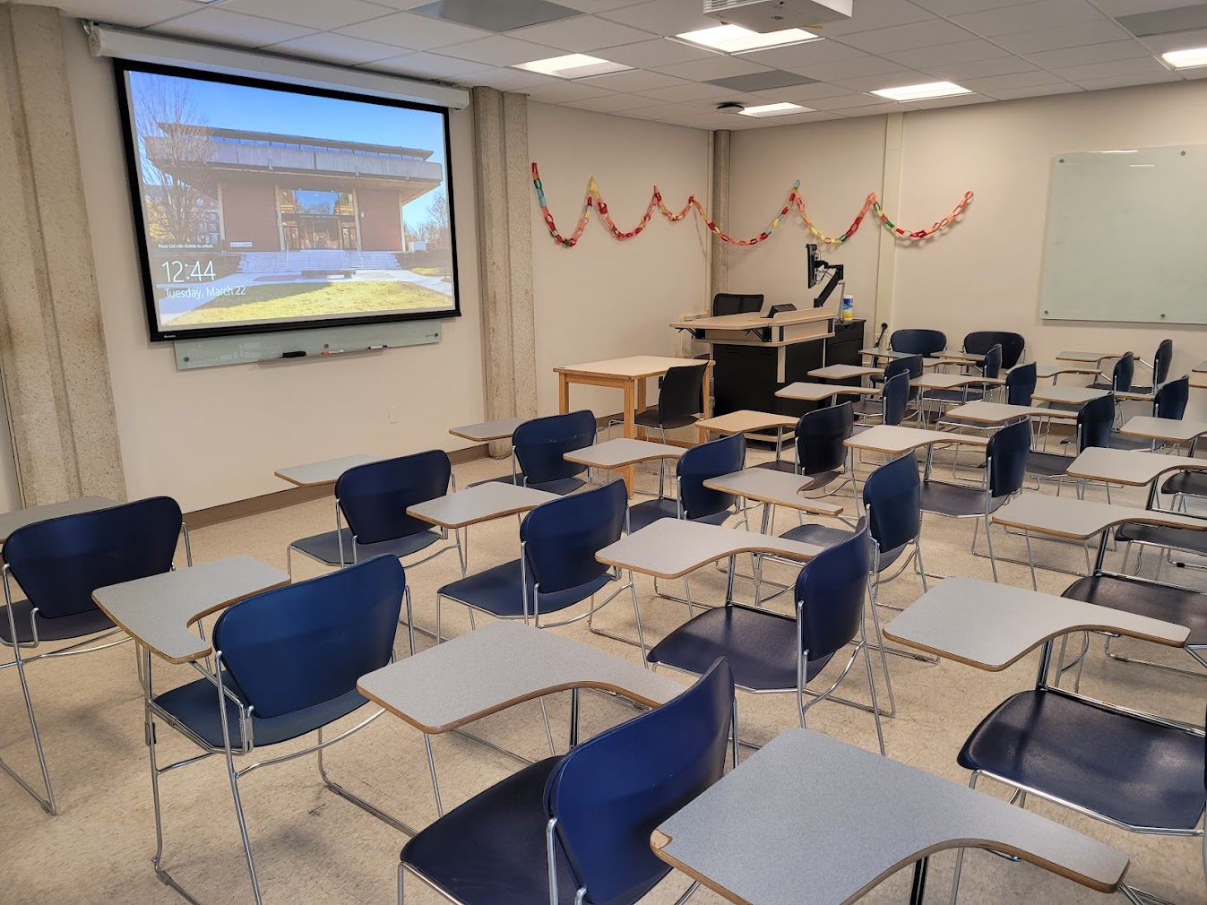 A view of the classroom with movable tableted arm chairs, glass board, and instructor table in front.