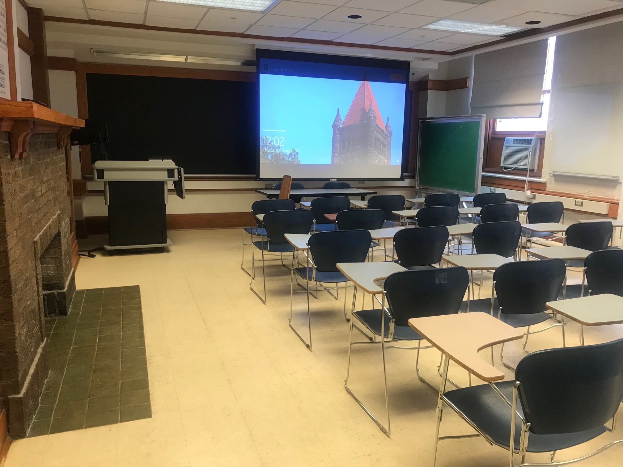A view of the classroom with movable tableted arm chairs, projector and screen, chalk board, instructor lectern, and instructor table in front.
