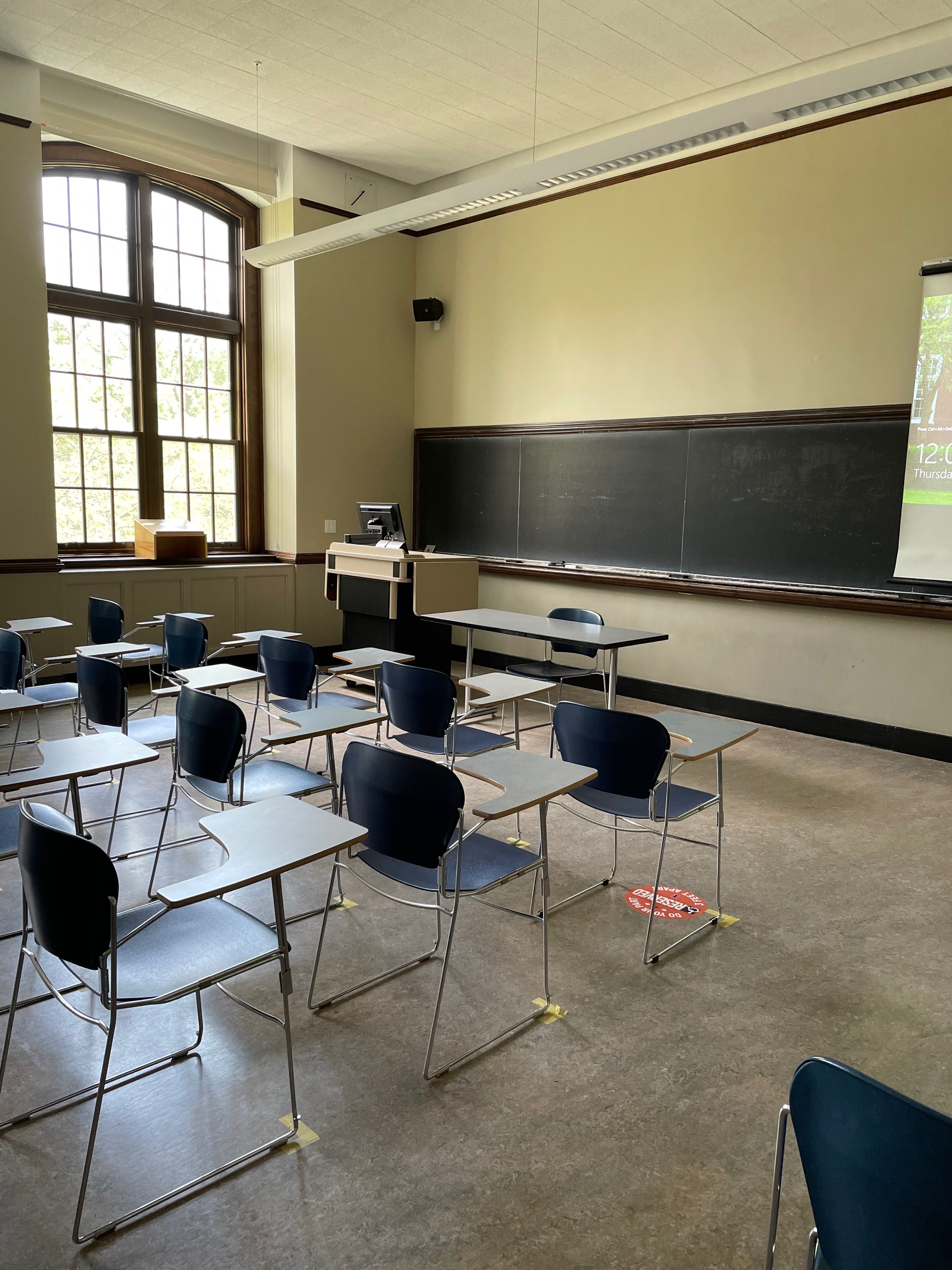 A view of the classroom with movable tableted arm chairs, chalkboard, and instructor table in front. 