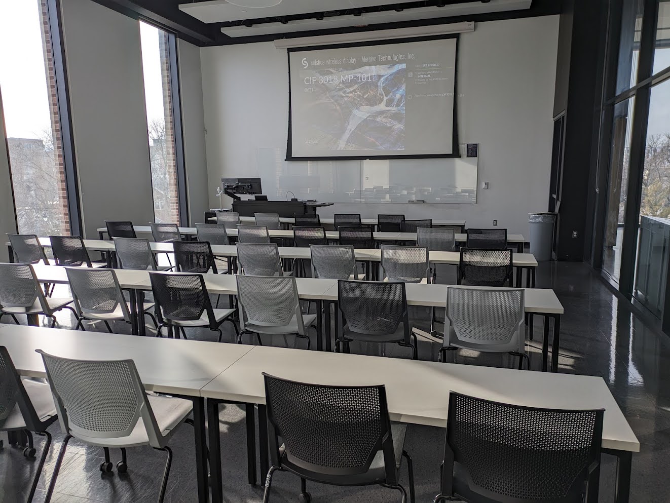 This is a view of the room, with student desks, a front lectern, projection screen, and a glass board.