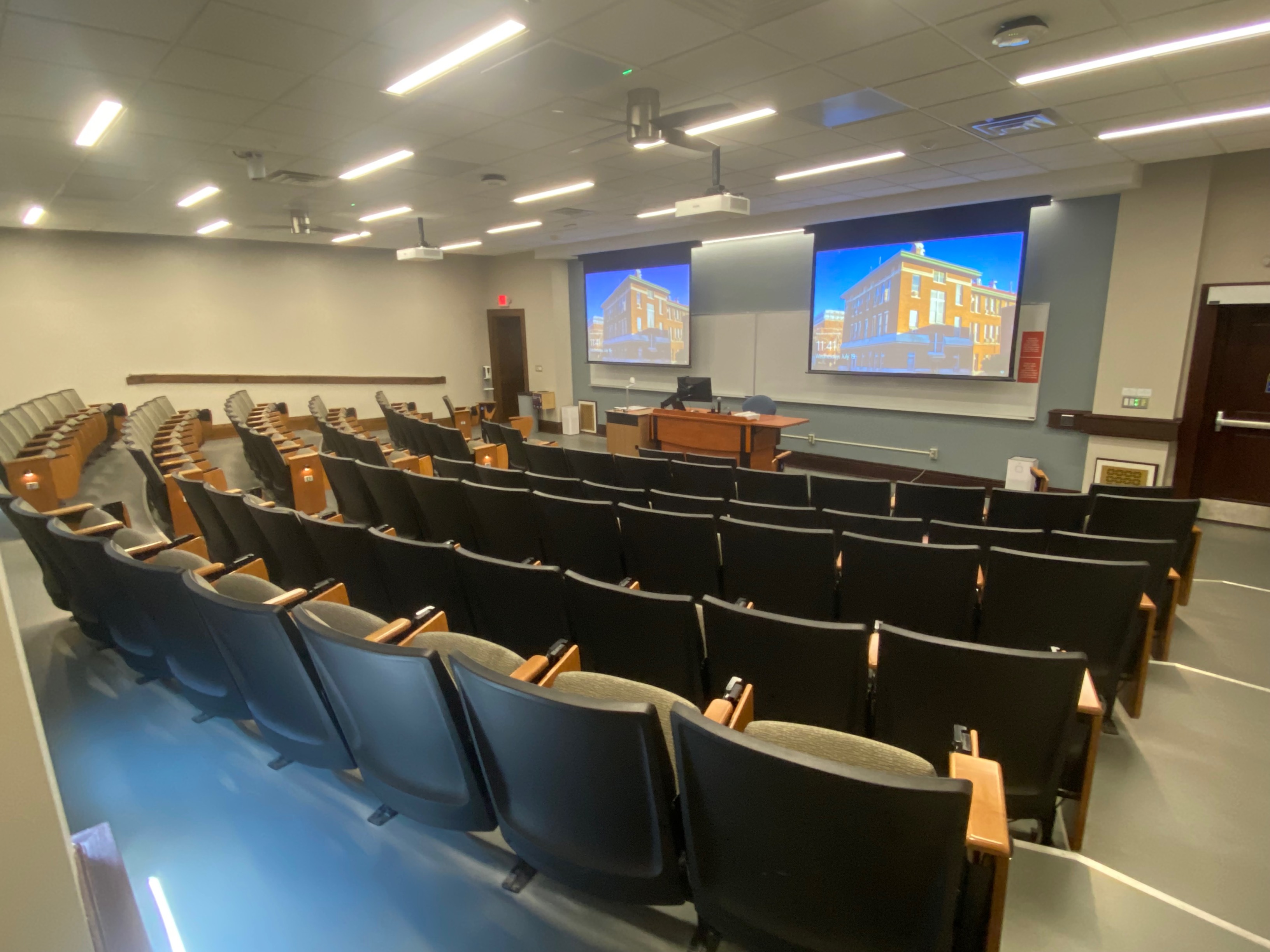 A view of the classroom with theater auditorium seating, projectors and screens, white boards, and instructor lectern in front. 