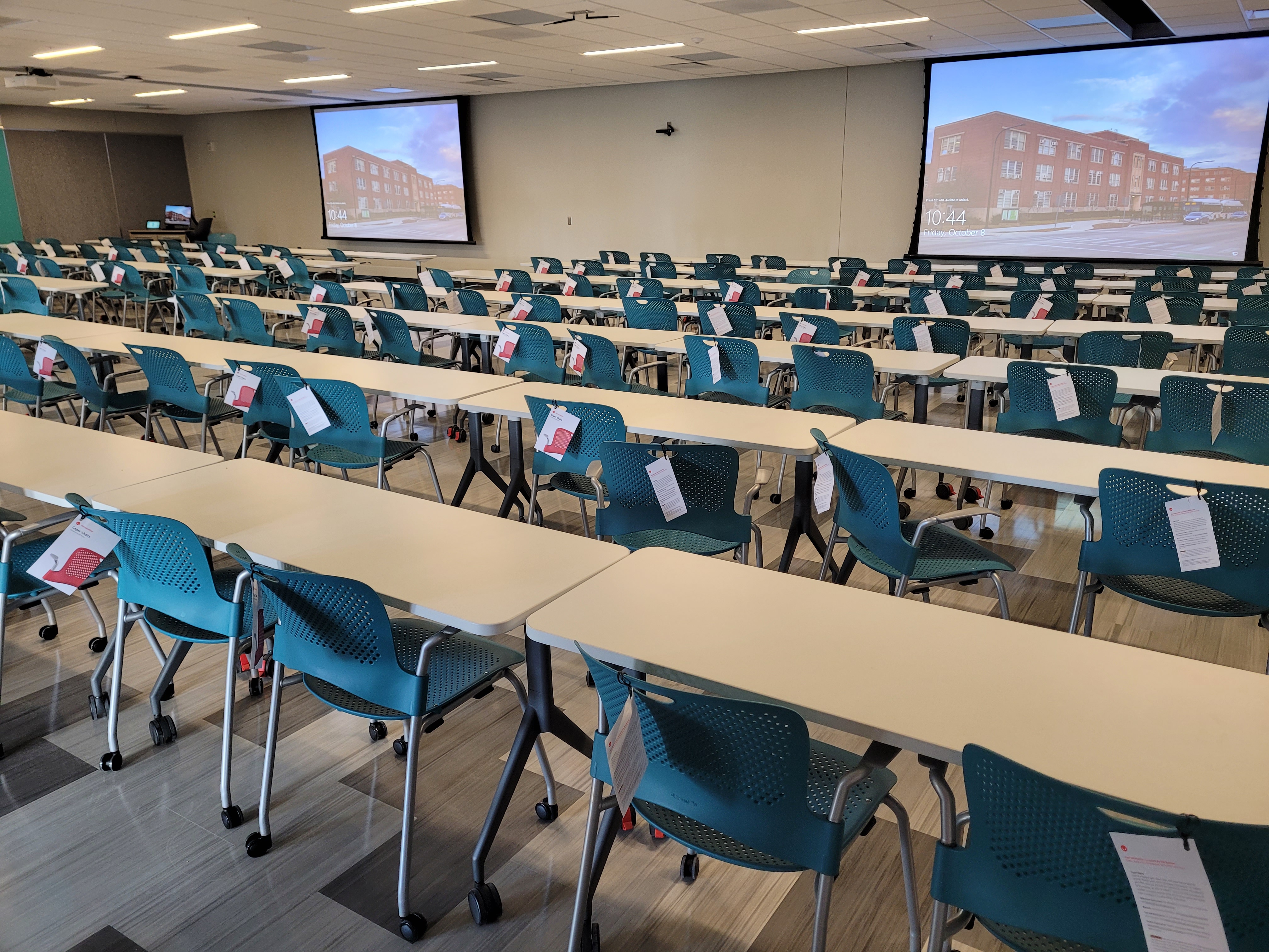 The view of the room with student tables and chairs in a row, a front lectern, and two projector screens.