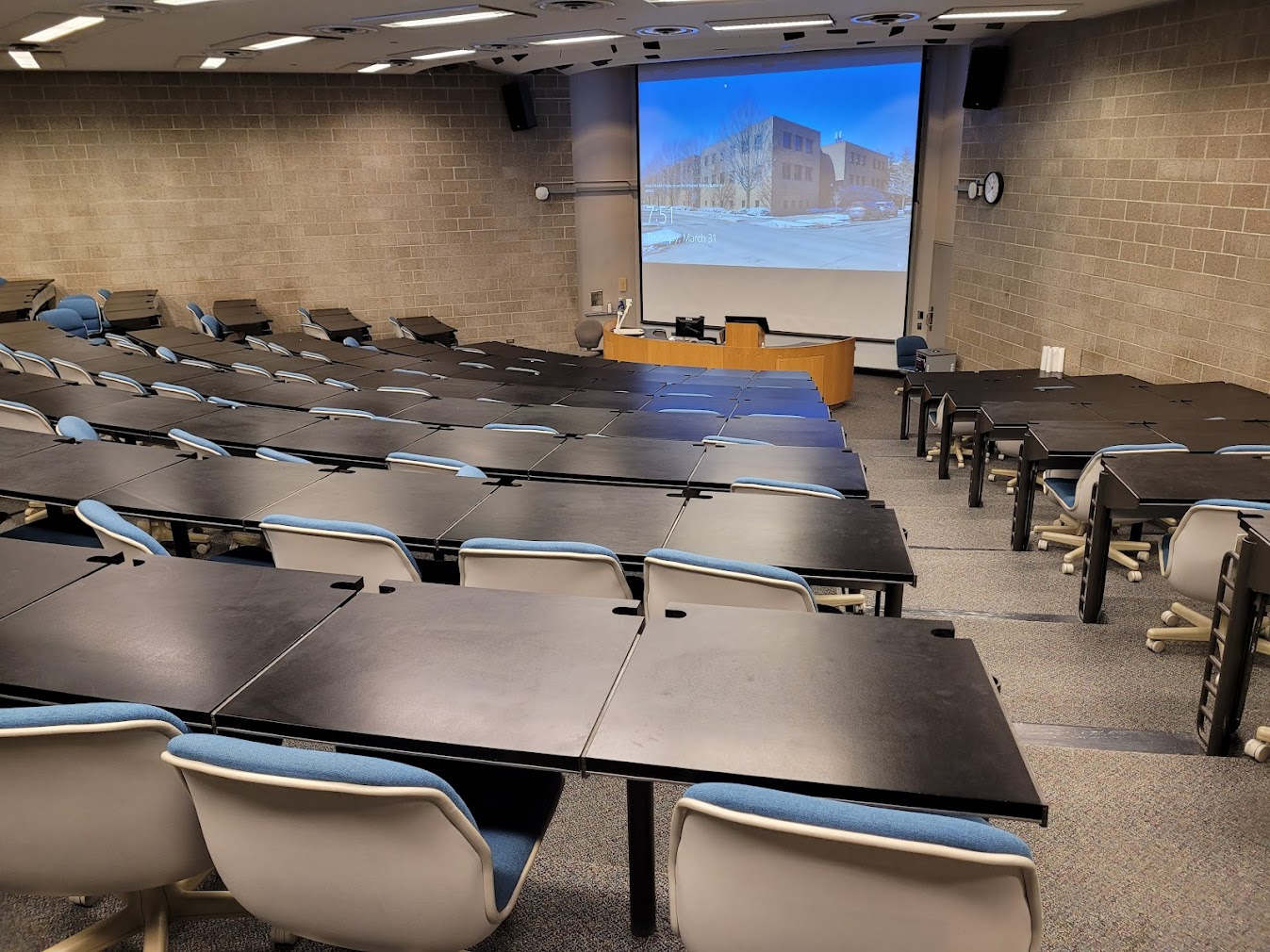 A view of the classroom with tiered, sloped seating, white board, and large lectern in front.
