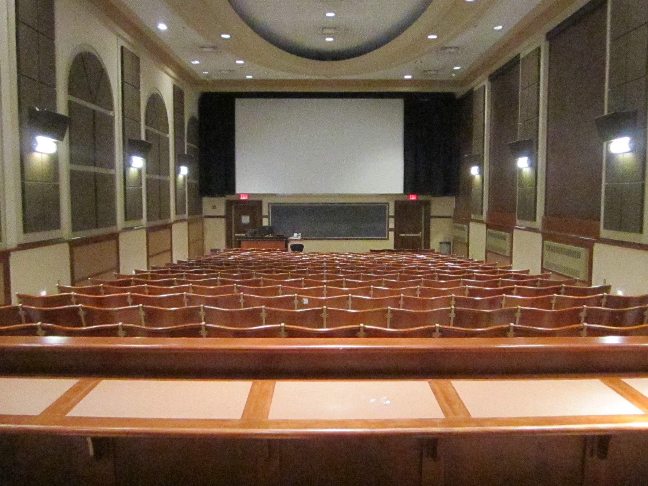 A view of the classroom with fixed auditorium seating, chalkboard, and projection screen in front.