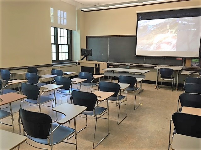 A view of the classroom with movable tableted arm chairs, chalkboard, and instructor table in front. 