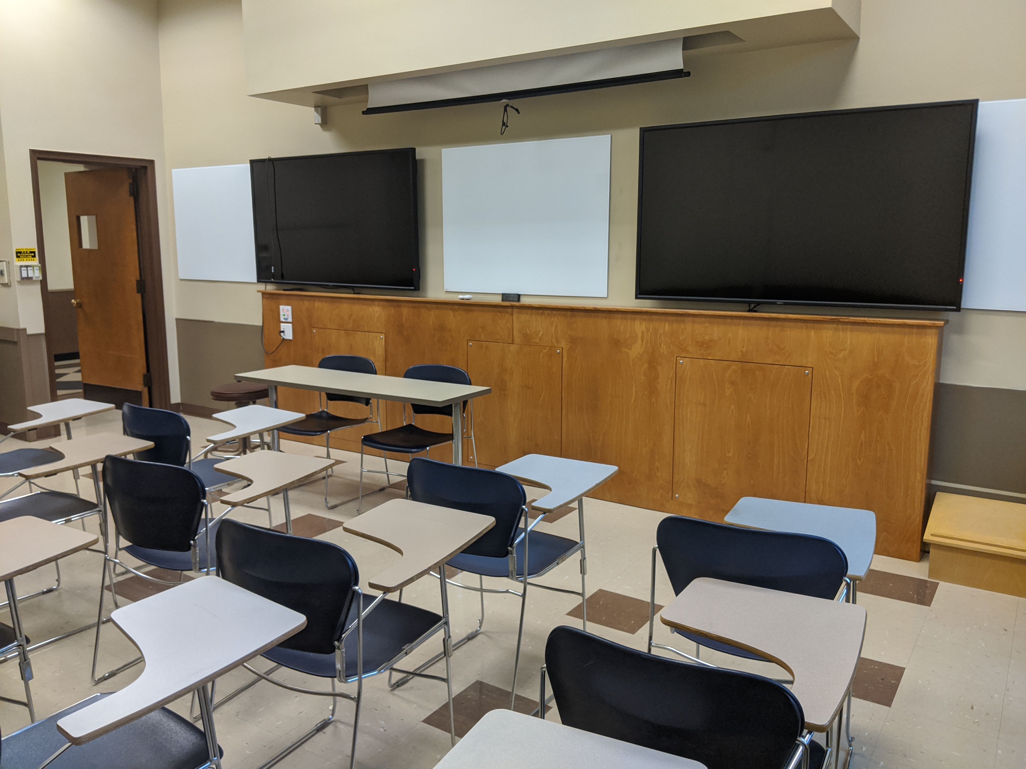 A view of the classroom with moveable tablet arm chairs, LCD wall monitors, whiteboards, and instructor table in front.