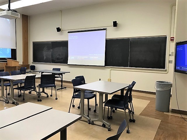 A view of the classroom with movable tables and chairs, TV monitors, and instructor table in front.