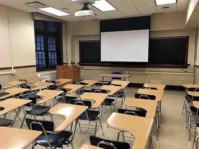 A view of the classroom with movable tables and chairs, chalkboard, and instructor table in front.