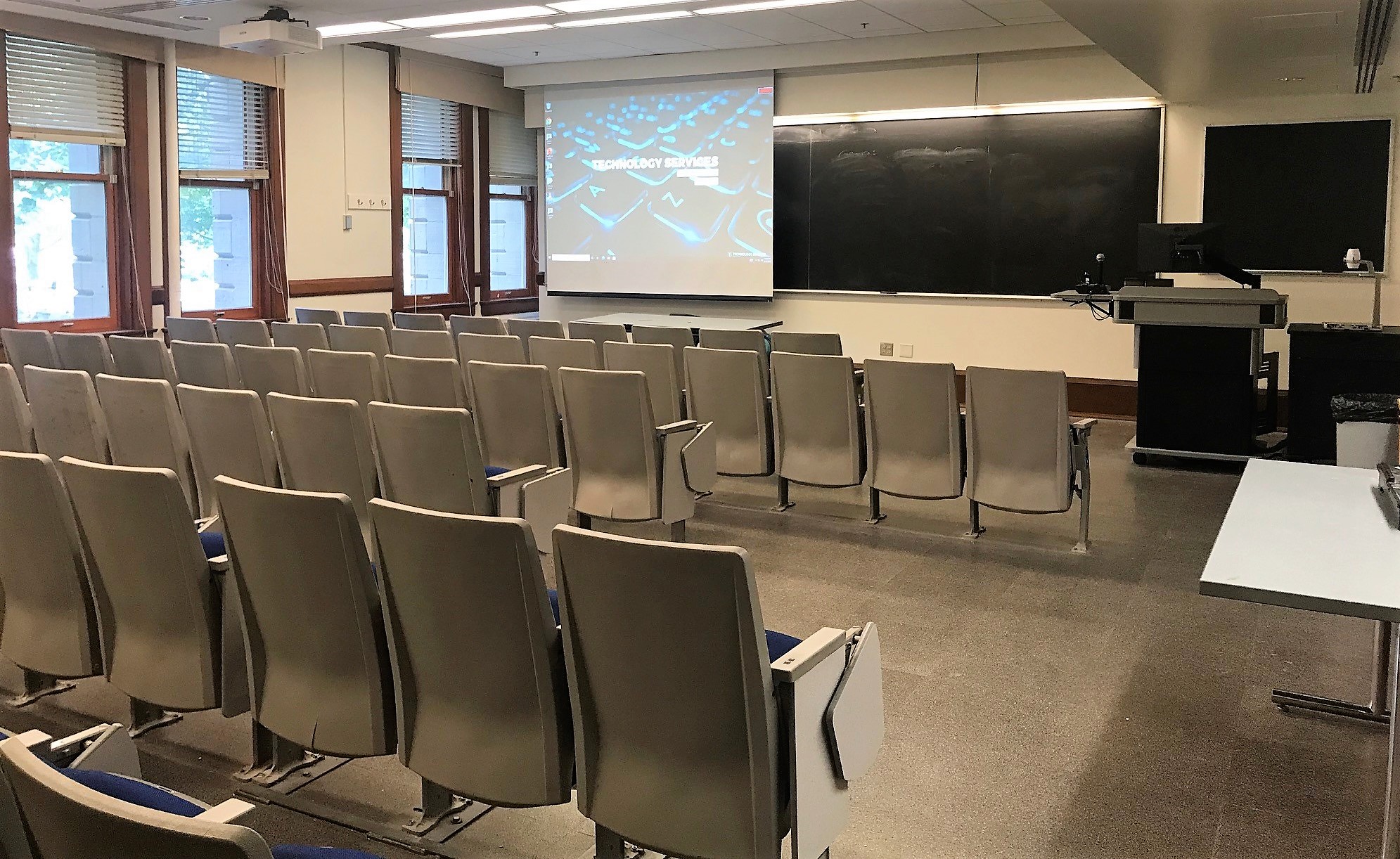 A view of the classroom with theater auditorium seating, chalkboard, projection screen, and instructor lectern in front.