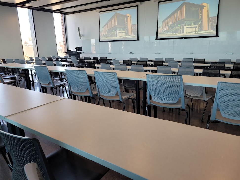 This is a view of the room with student desks, a front lecture table, windows, and glass boards.
