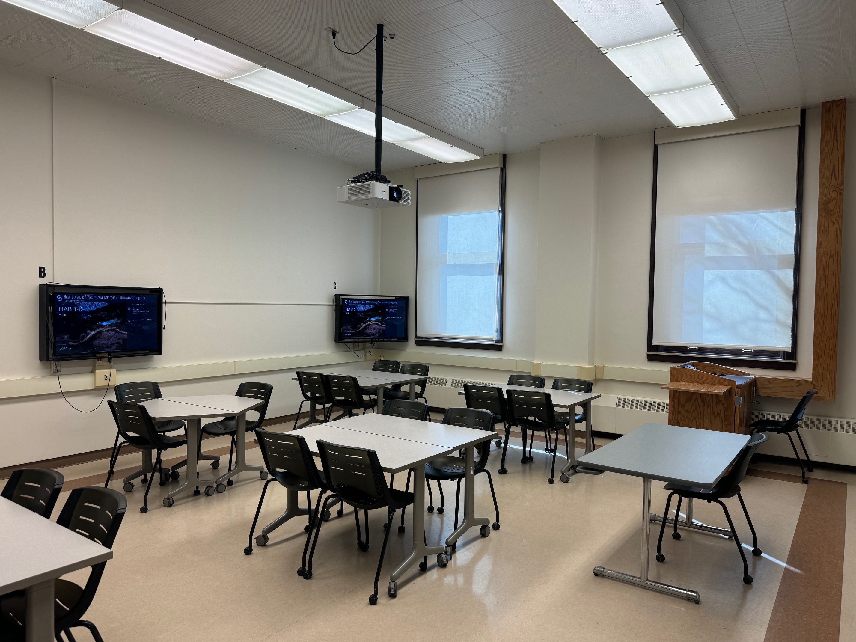 A view of the classroom with movable tables and chairs, tv monitors, and instructor table in front.