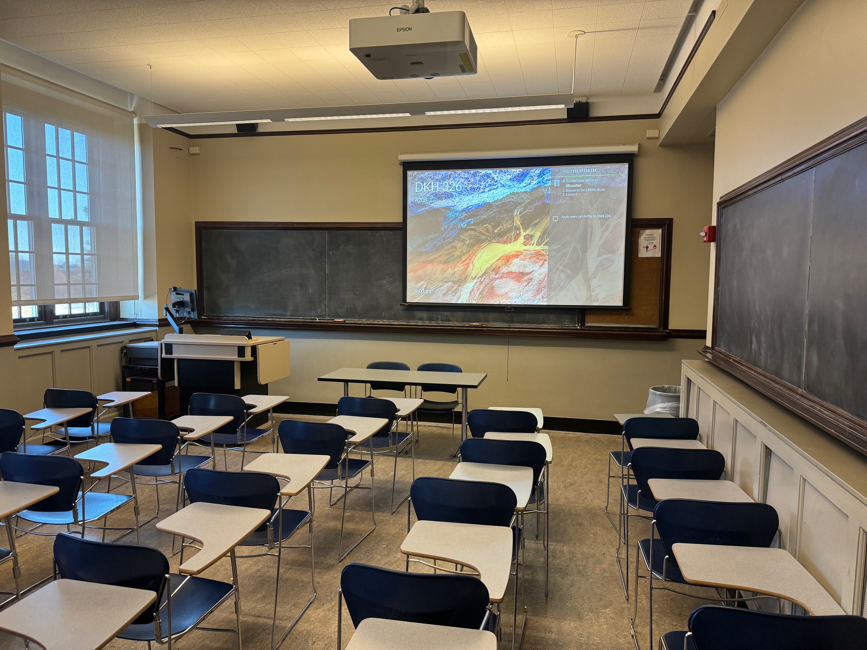 A view of the classroom with tiered sloped seating, chalkboard, and instructor table in front.