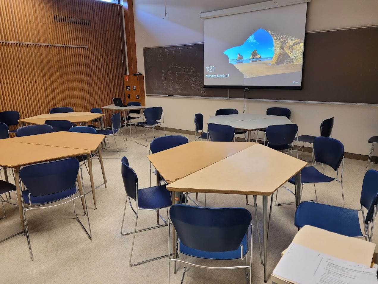 A view of the classroom with moveable tables and chairs, projection screen, and chalk board.